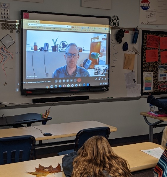 Classroom setting with a presenter on a screen in front of the room.