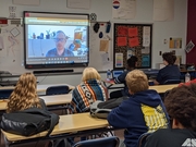 Classroom setting with a presenter on a screen in front of the room, some students facing screen sitting at their desks.
